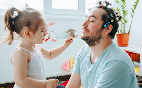 Girl adding lip gloss to adoptive dad. He has his eyes closed and also has hair clips in his hair.